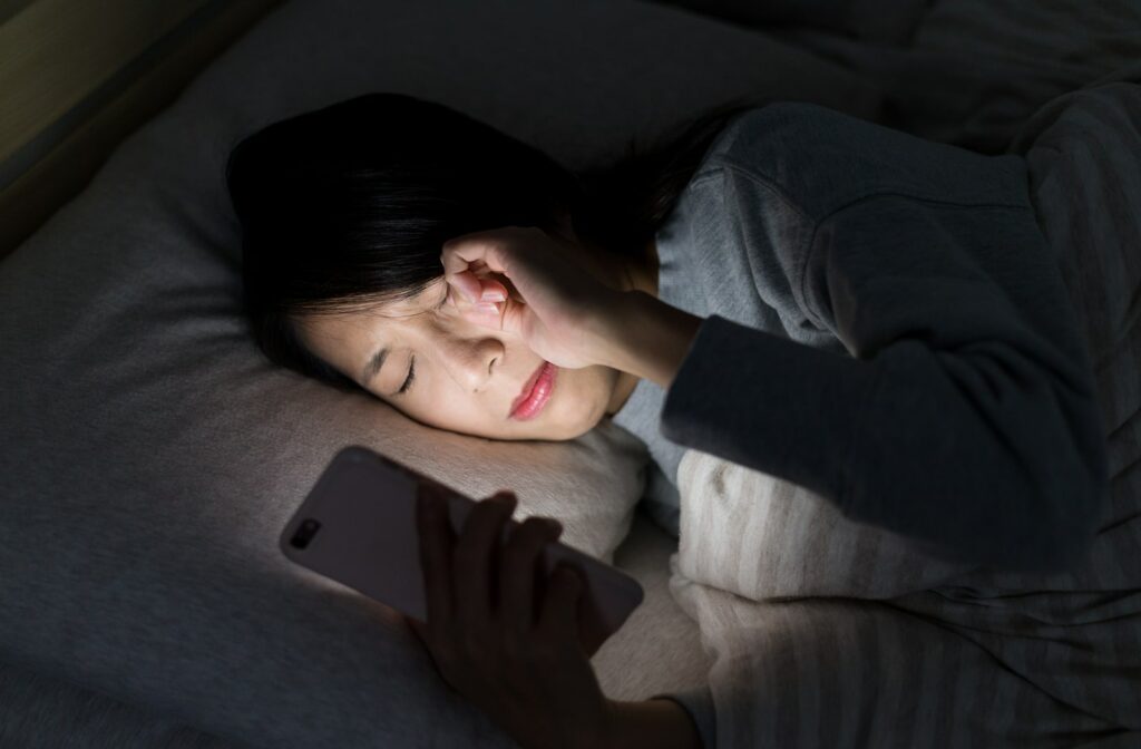 A woman rubbing her dry eyes while looking at her phone in bed