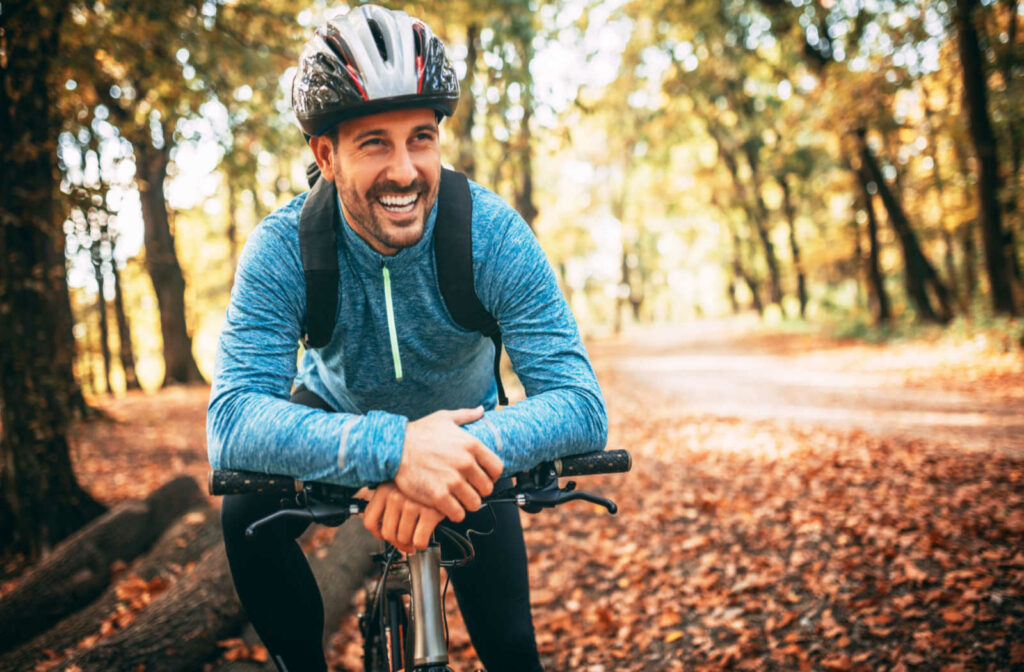 A smiling bicyclist leaning over the handlebars surrounded by trees and fallen leaves.