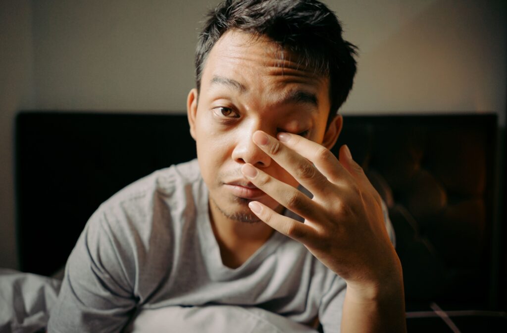 Young man rubbing left eye with finger while sitting up from bed.