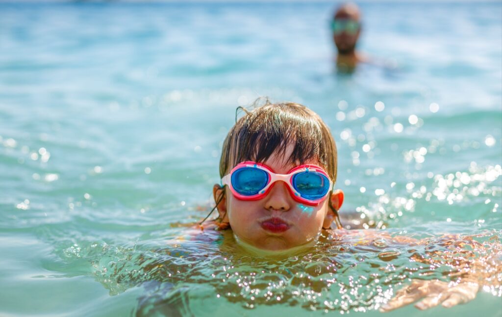 A child wearing prescription swim goggles paddles through some ocean water on a hot summer day.