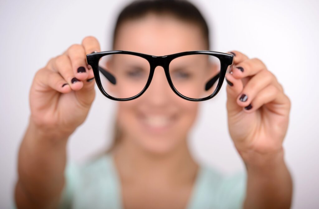 A close-up of a black pair of glasses held by a blurred figure in the background, symbolizing the need for corrective lenses.